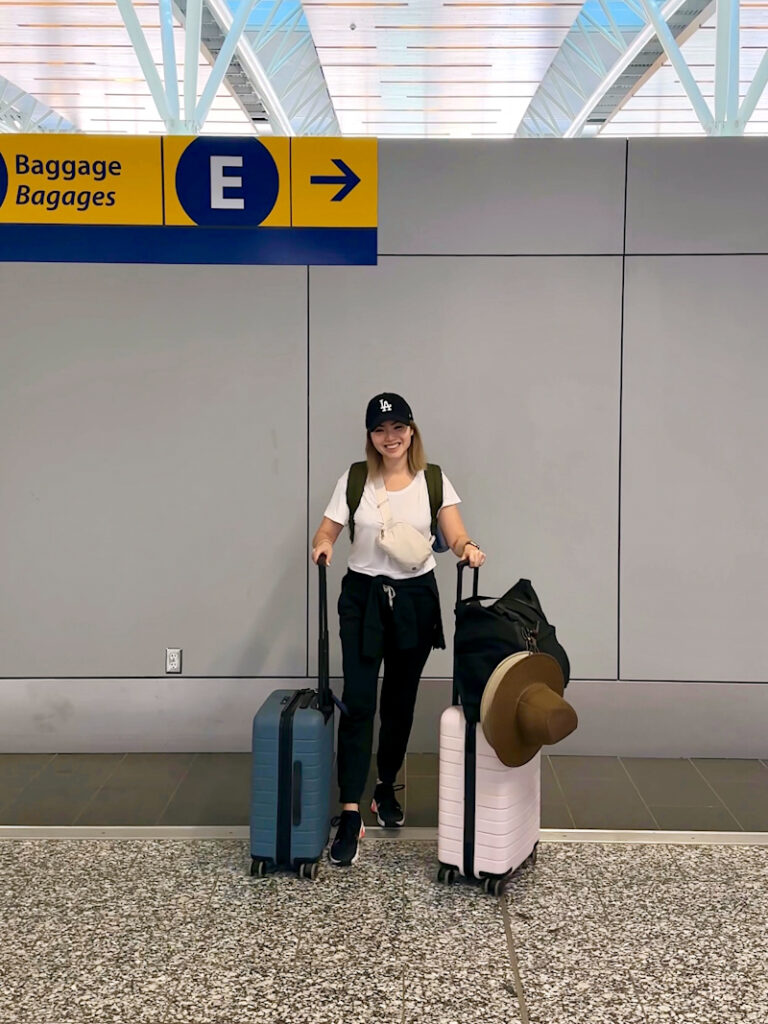 Girl in airport packed for international trip with carry on travel only. She's holding two Away luggages, a black duffel bag, and a hat.
